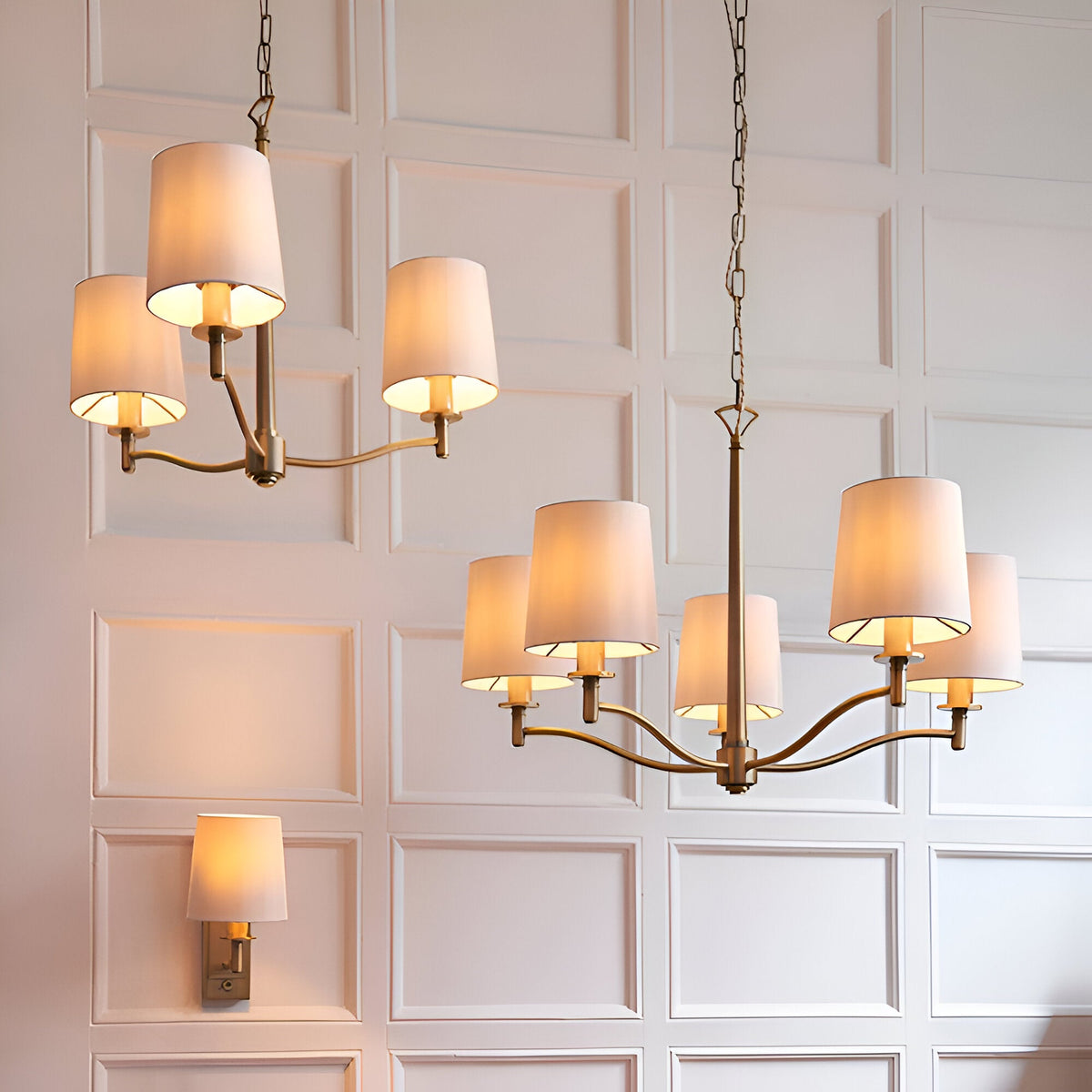 Endon Castwell 5-light pendant in antique brass with vintage white fabric shades, shown with matching 3-light pendant and wall light on a panelled wall.