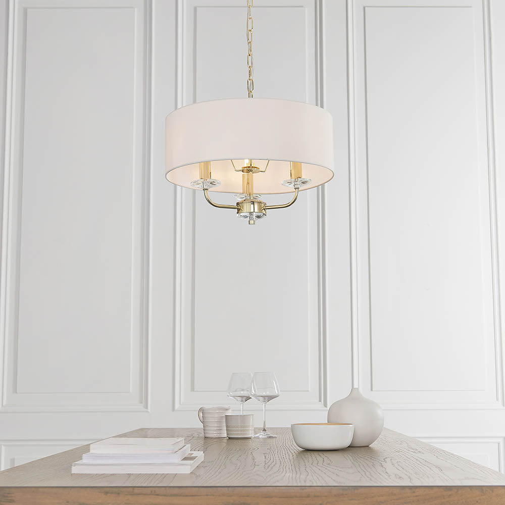 Endon Castdon 3 light pendant in brass with white faux silk drum shade and crystal detail, hanging above a wooden dining table against a panelled wall.