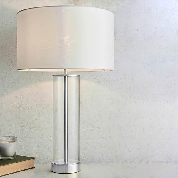 Endon Caldley table lamp with a clear cylindrical glass base, nickel fittings and white drum shade, styled on a console beside a candle and book.