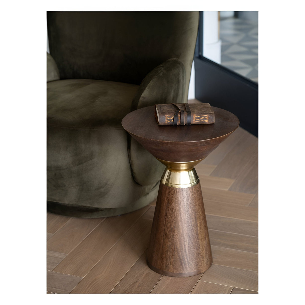 Libra Interiors Amalfi side table in dark mango wood with brass-effect band, beside a green velvet armchair, with a leather-bound journal on top.