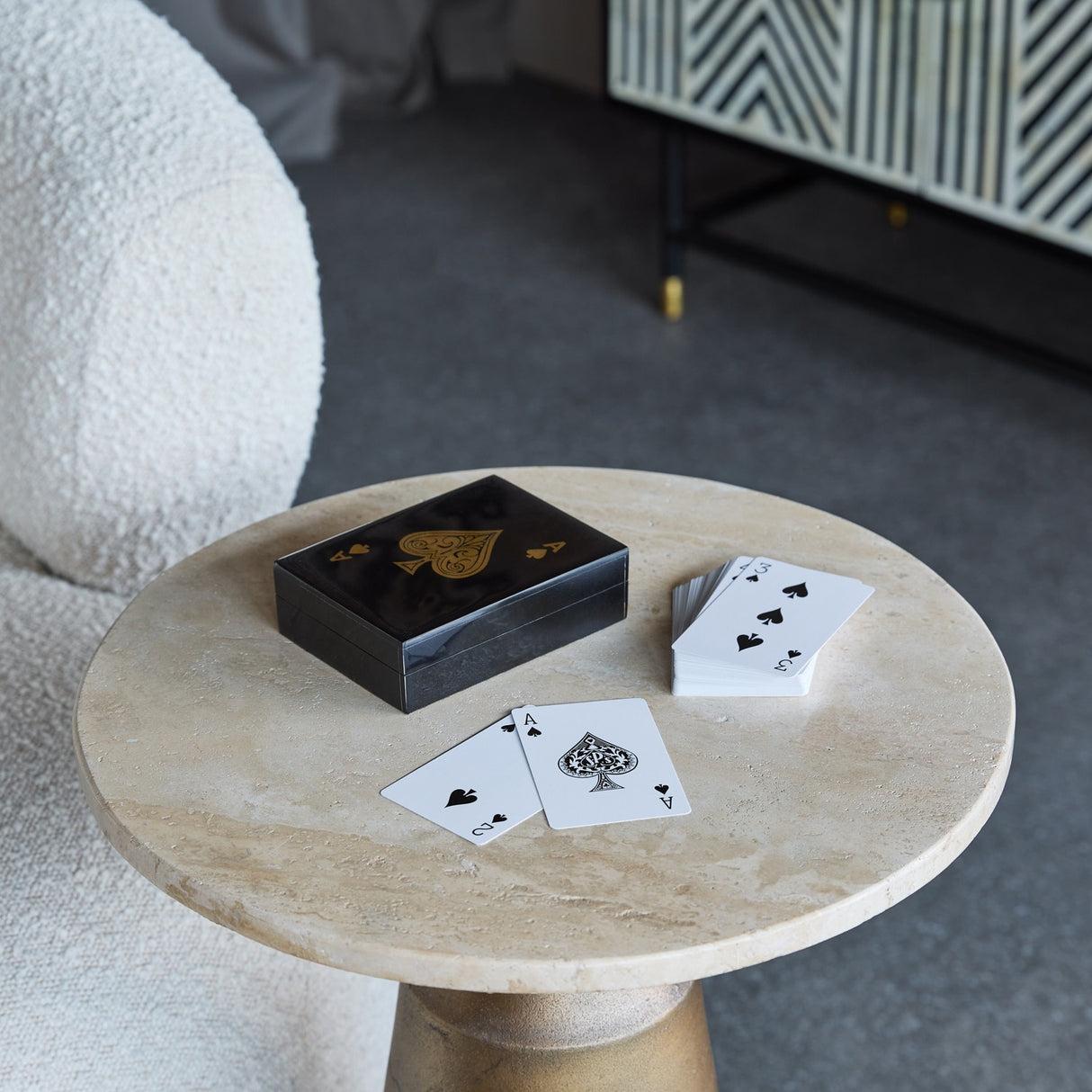 Libra Interiors Ace of Spades black resin card game box with gold motif on a round travertine side table, deck fanned beside it in a living room.