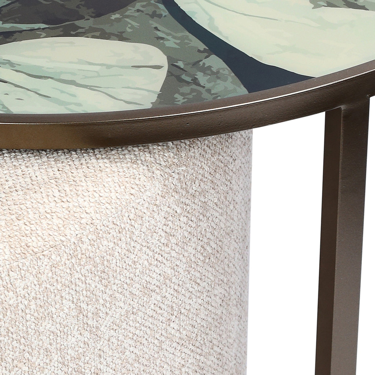 Close-up of Libra Interiors Printed Glass Nesting Coffee Table with muted green leaf-printed top, bronze-tone iron frame and oatmeal fabric pouffe beneath.