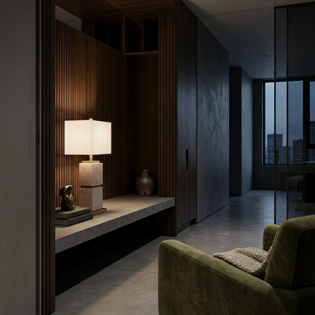 A modern, dimly lit hallway with wood paneling features a concrete bench holding the Veradex 1 Light Marble Block Table Lamp in Brass & Purple, books, a small sculpture, and a vase. An olive green armchair and large city-view windows are in the background.