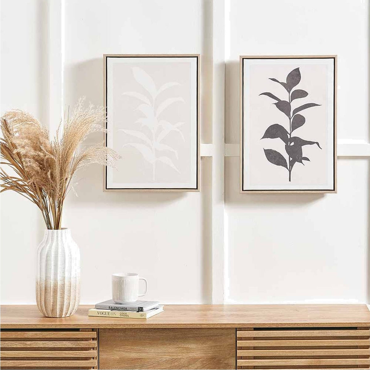 Set of 2 framed leaf print canvases, one natural and one black on light background, hung above a wooden sideboard with a vase and books.