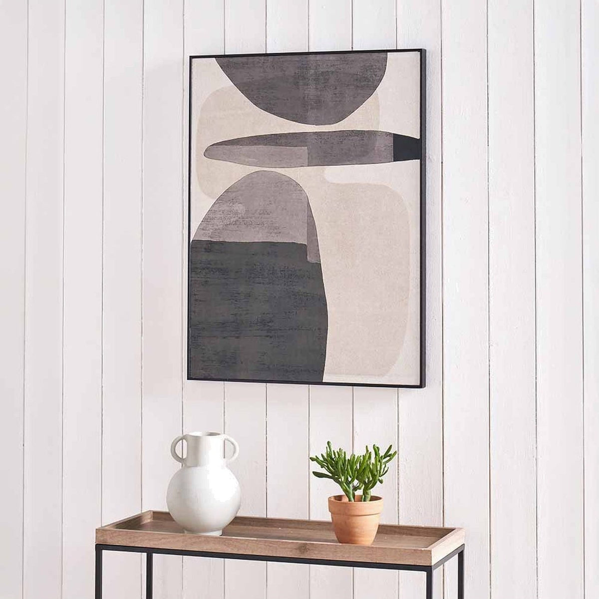 Framed canvas wall art in black and natural tones featuring abstract oval shapes, hung above a wooden console table with a white vase and potted plant.