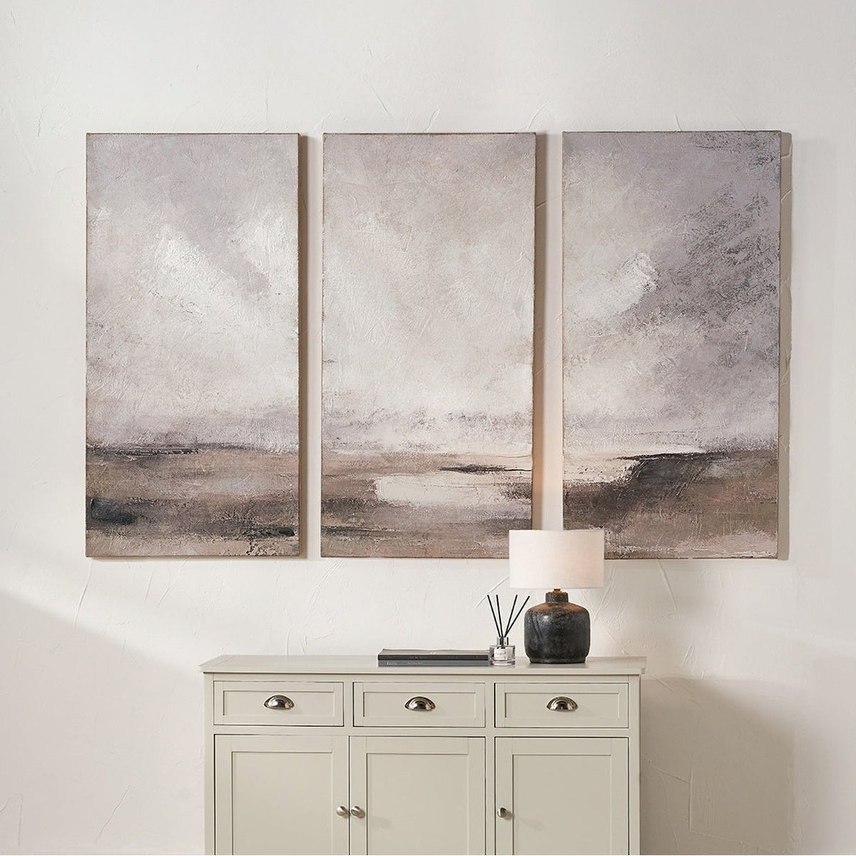 Set of three grey and neutral abstract landscape canvas wall art panels above a light cabinet, each canvas showing soft cloud-like tones over a darker horizon band.