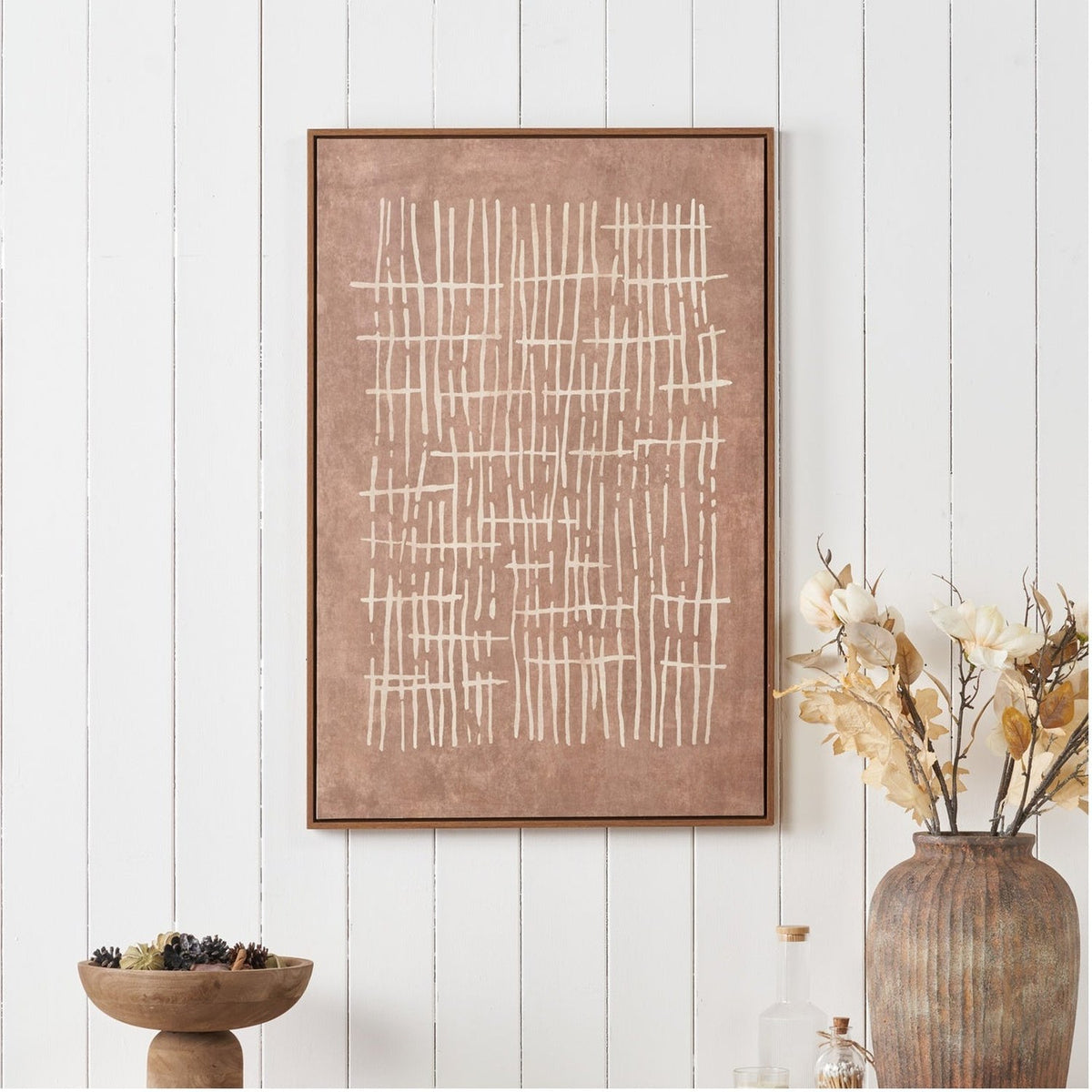 Canvas wall art with abstract cream linear crosshatch design on a brown background, framed in oak-effect wood, displayed on a white panelled wall above a wooden bowl and vase.