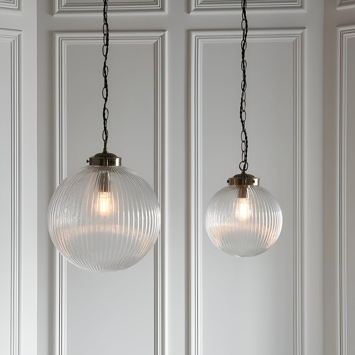 Endon Ashmere pendant lights in two sizes with ribbed clear glass globe shades and antique brass chains, hung against a white panelled wall.