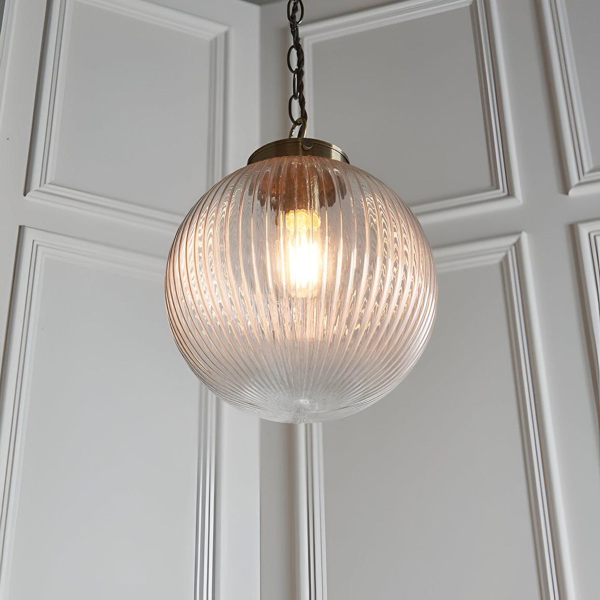 Endon Ashmere pendant light with ribbed round clear glass globe shade and antique brass chain suspension against white panelled wall.