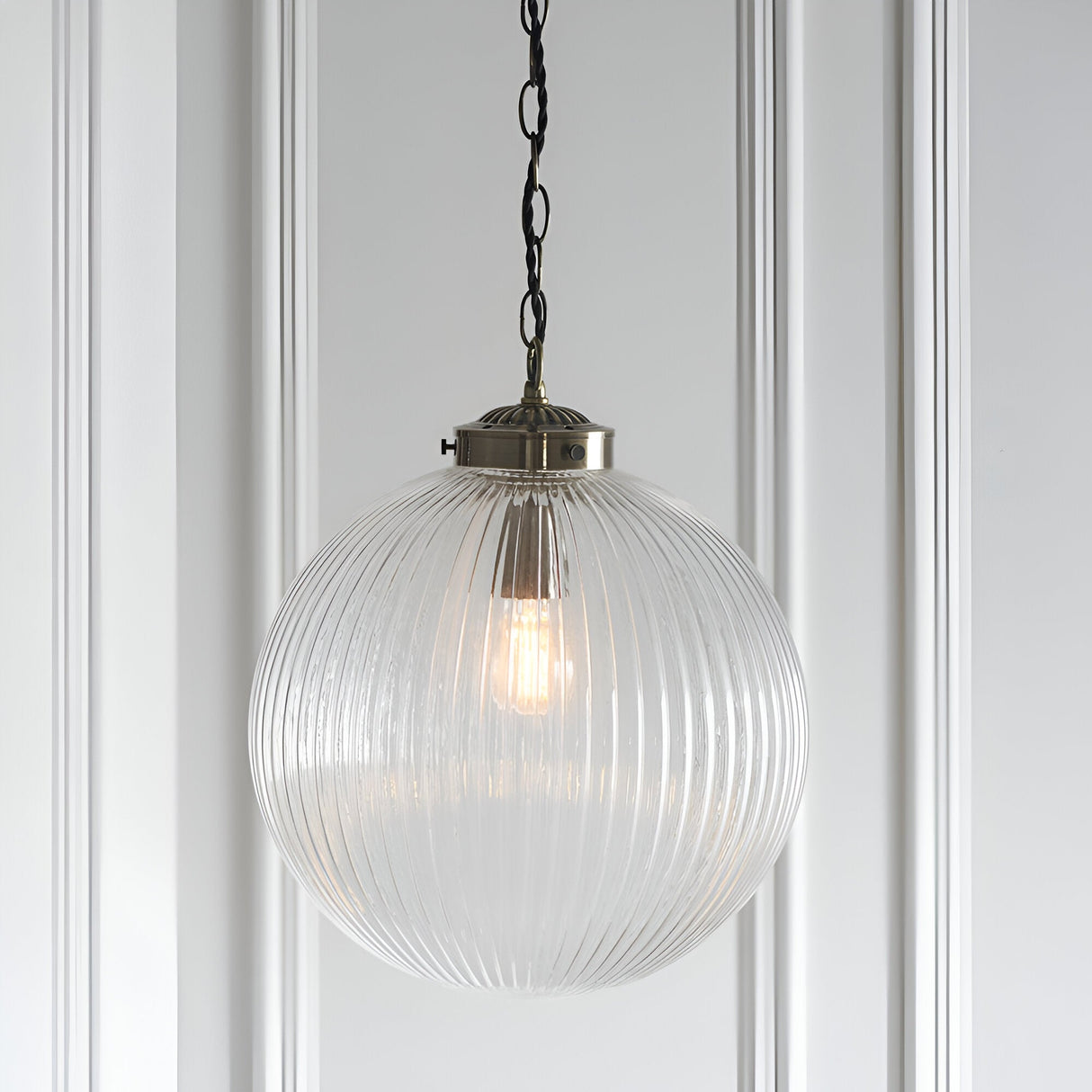 Endon Ashmere Large pendant light with ribbed round clear glass globe shade and antique brass chain suspension, mounted against a white panelled wall.