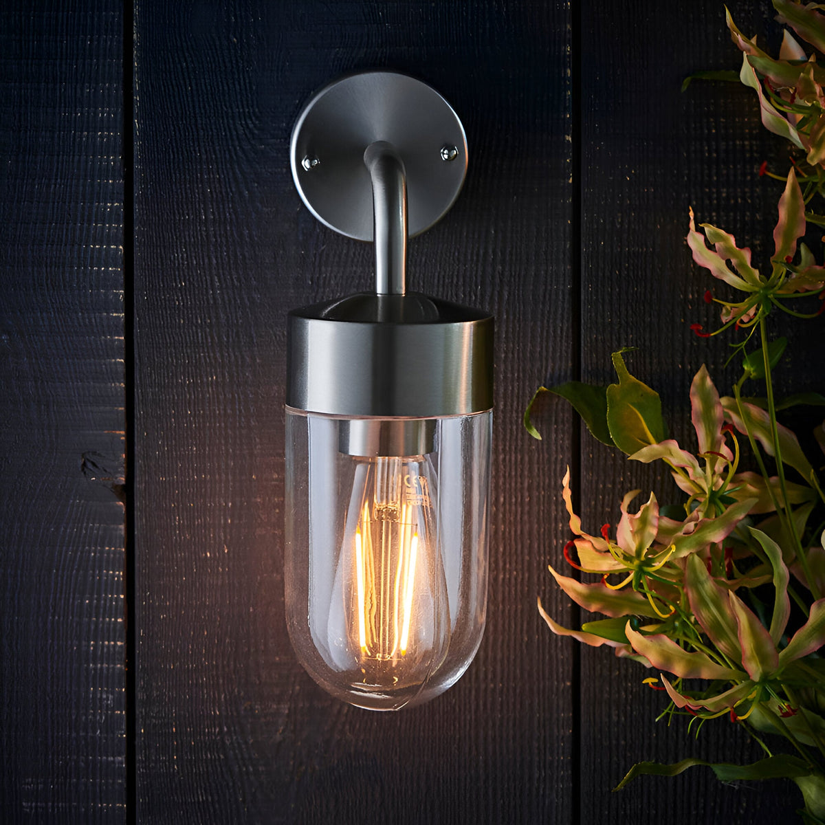 Endon Castfield outdoor wall light in silver steel with clear glass cylinder shade and exposed E27 filament bulb, mounted on a dark timber wall beside foliage.