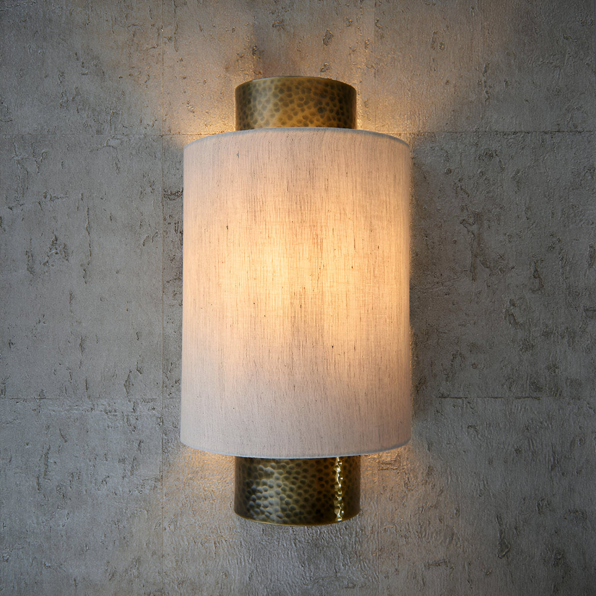 Endon Brenwood wall light with hammered aged bronze fittings and a natural linen-effect fabric shade, illuminated against a textured grey concrete wall.