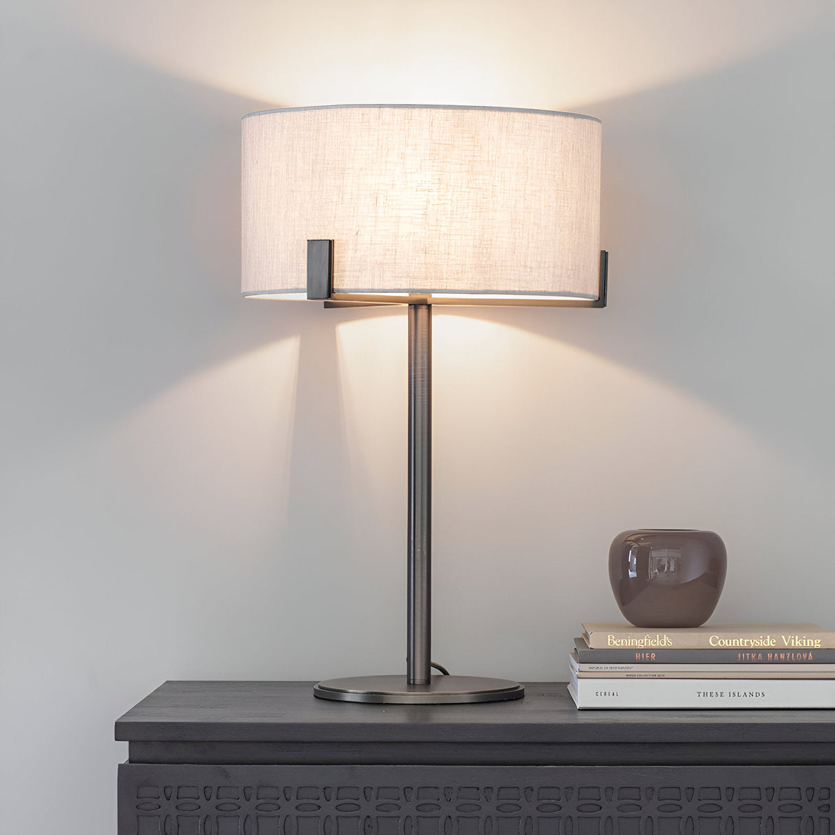 Brendon Table Lamp in bronze finish with a natural linen drum shade, shown lit on a dark sideboard beside books and a decorative vase.