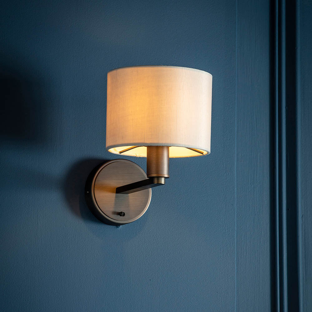 Endon Bexley wall light illuminated on a deep blue panelled wall, with a bronze circular backplate and arm supporting a cream faux silk drum shade.