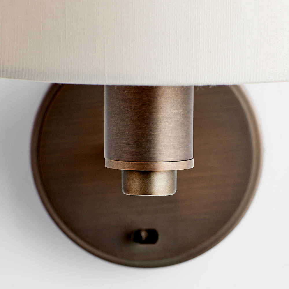 Close-up of the Endon Bexley wall light showing the dark antique bronze cylindrical arm, circular backplate with fixing screw, and base of the faux silk shade.
