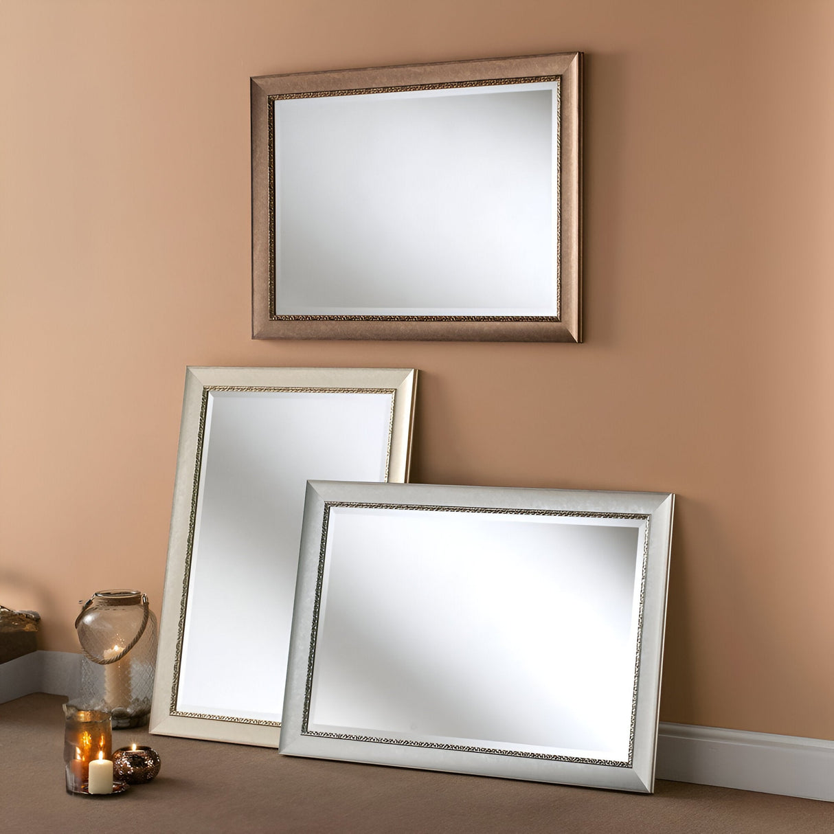 Three Zeal Rectangle Wall Mirrors in a decorative champagne finish are positioned against a beige wall. Two mirrors rest on the carpeted floor, while one is hung above them. A lit candle and a decorative lantern complete this cozy home decor scene, adding warmth and charm to the setting.