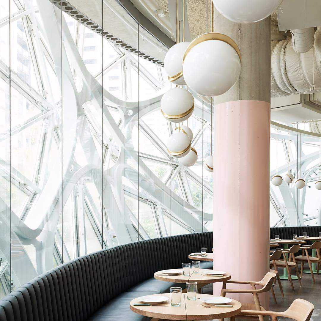 A modern, airy restaurant featuring large geometric glass windows and round wooden tables with stylish place settings, complemented by curved black seating. The space is enhanced by the contemporary design of Arc Pendant Lights in gold and white and brushed brass accents.