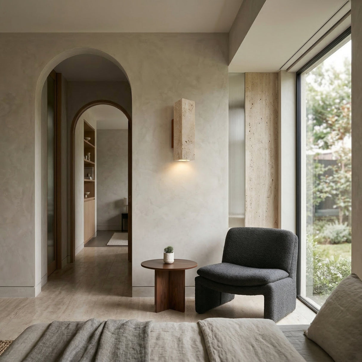 A modern, minimalist living space with a dark gray armchair, walnut brown round side table with a potted plant, large garden-view window, light-toned walls, and a Bravora 1 Light Large Rectangle Travertine Wall Light in walnut brown & light beige by an arched doorway.