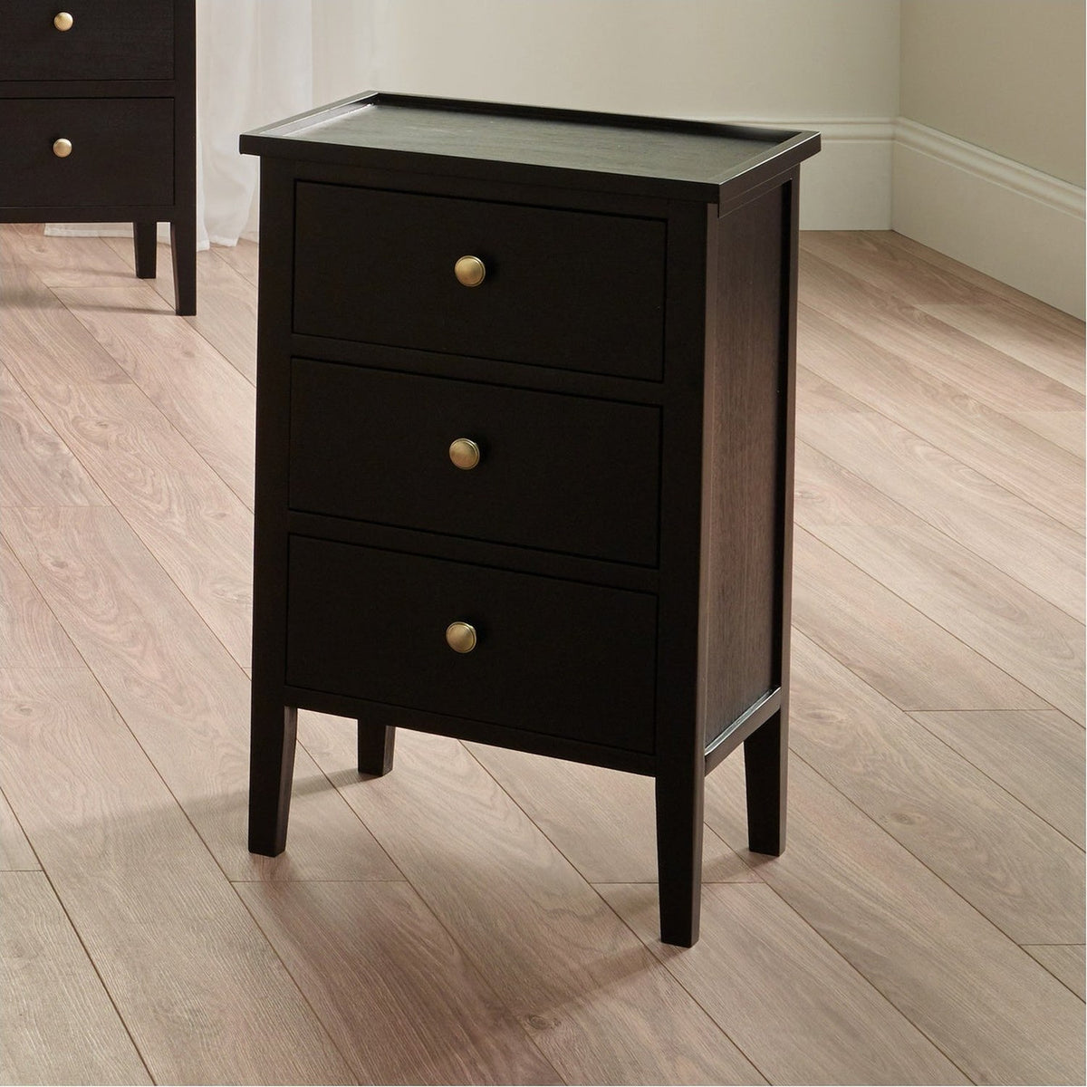 Chelmsford satin black pine 3 drawer unit with a raised-edge top and round brass knobs, shown in a bedroom setting on a wooden floor.
