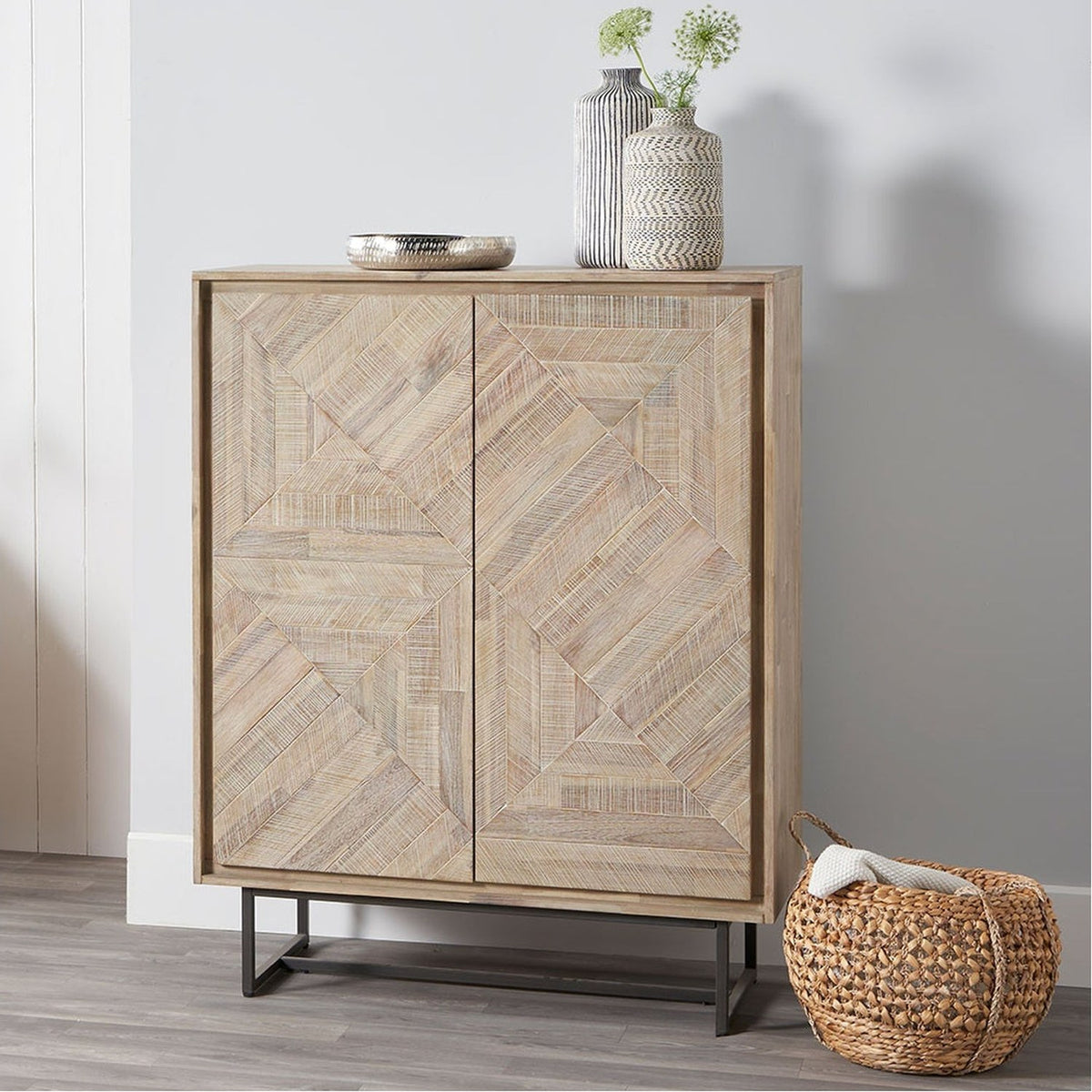 Marca sand wash acacia 2-door bar cabinet with geometric parquet-style doors and black iron legs, shown in a room with decorative vases on top.