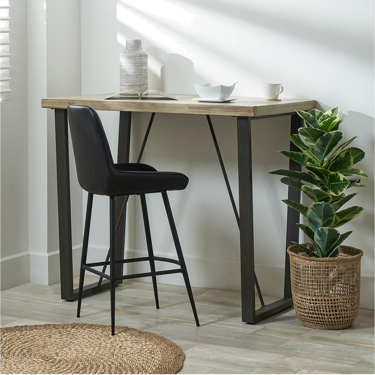 Marca Sand Wash Acacia & Black Iron Bar Dining Table, 107cm, with wood top and angled black metal legs, styled with a black bar stool, decor, and plant in a room setting.
