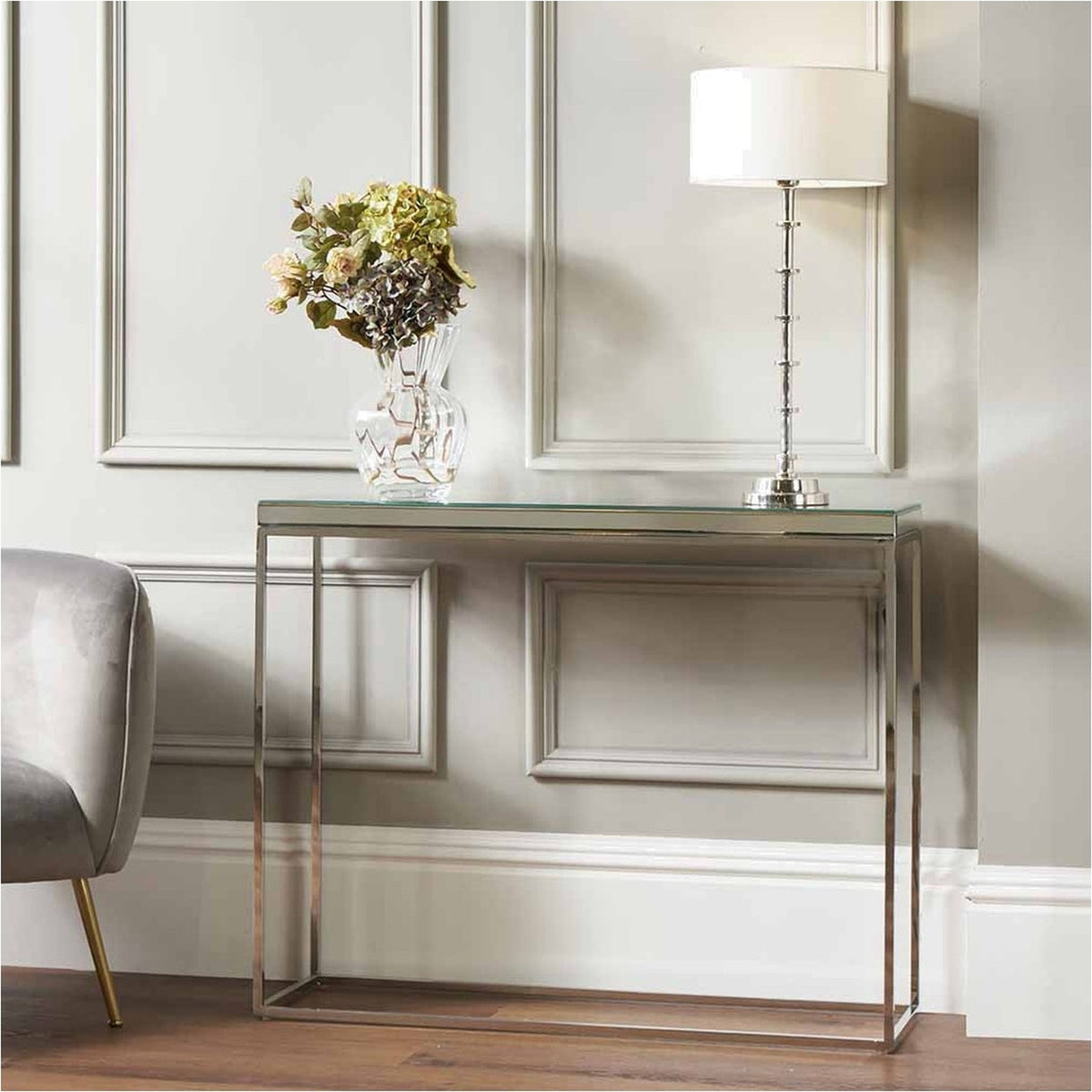 Elysee Mirrored Glass & Silver Metal Console Table with mirrored top and slim steel frame, styled with a vase of flowers and table lamp against a panelled wall.