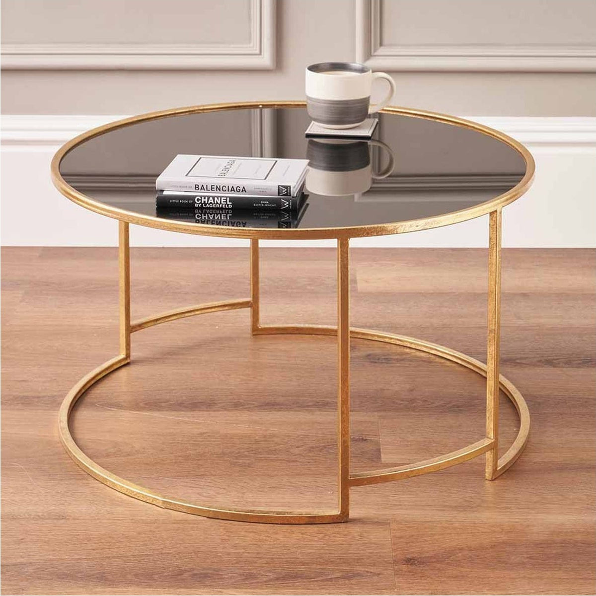 Round coffee table with black glass top and antique gold iron frame, double circular base, styled with books and a mug on a wooden floor.