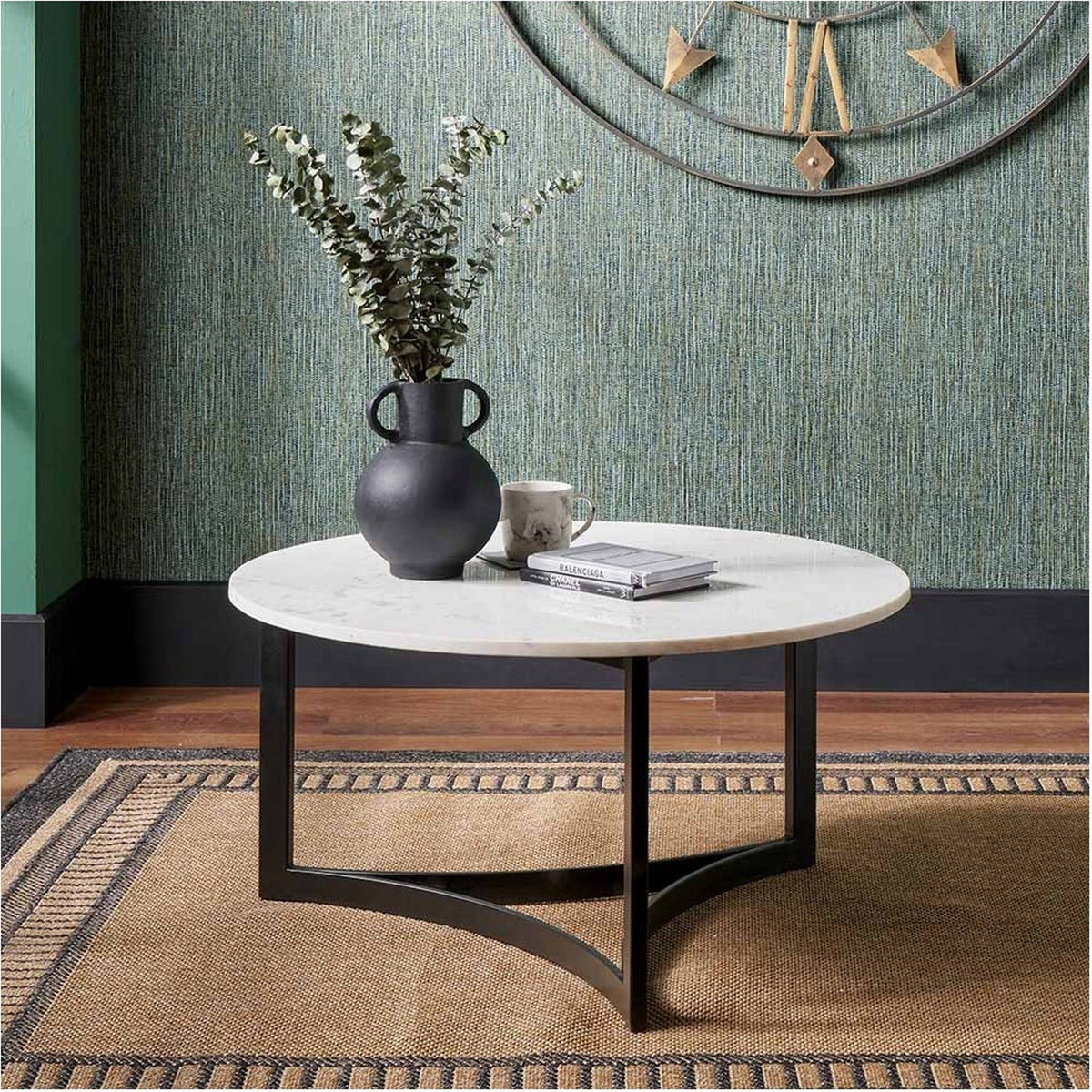 Hendrick round coffee table with white marble top and black curved iron base, styled in a living room with a black vase, plant, mug, and books on top.