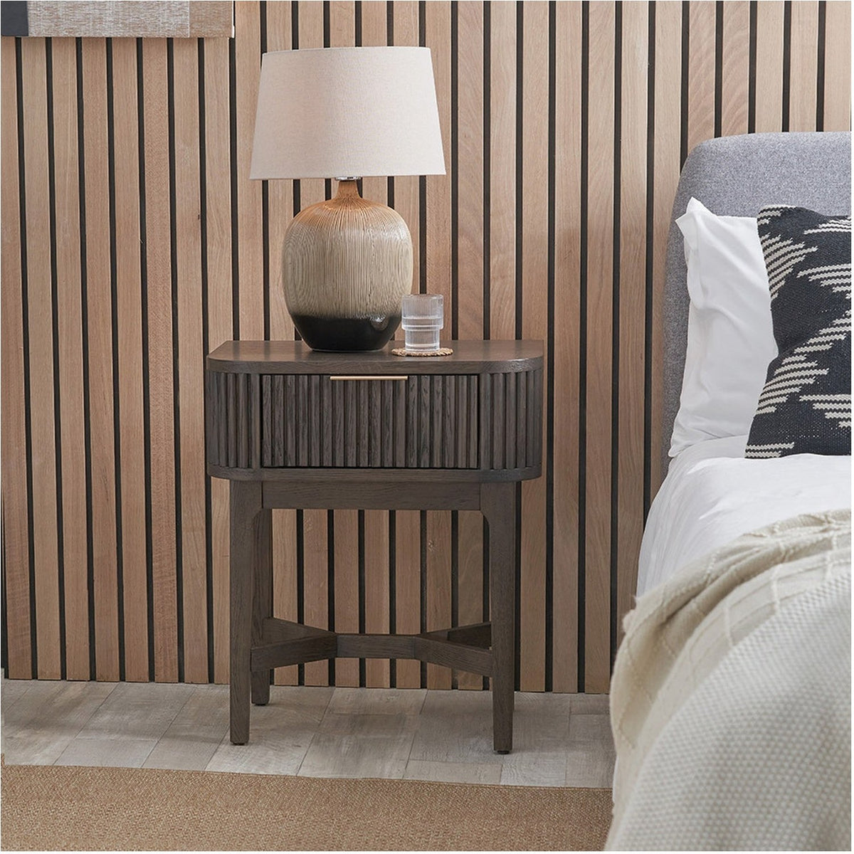 Raie Dark Brown Ribbed Oak & Pine 1 Drawer Unit used as a bedside table, with rounded top, ribbed front, brass handle, and splayed legs, styled beside a bed with a table lamp on top.