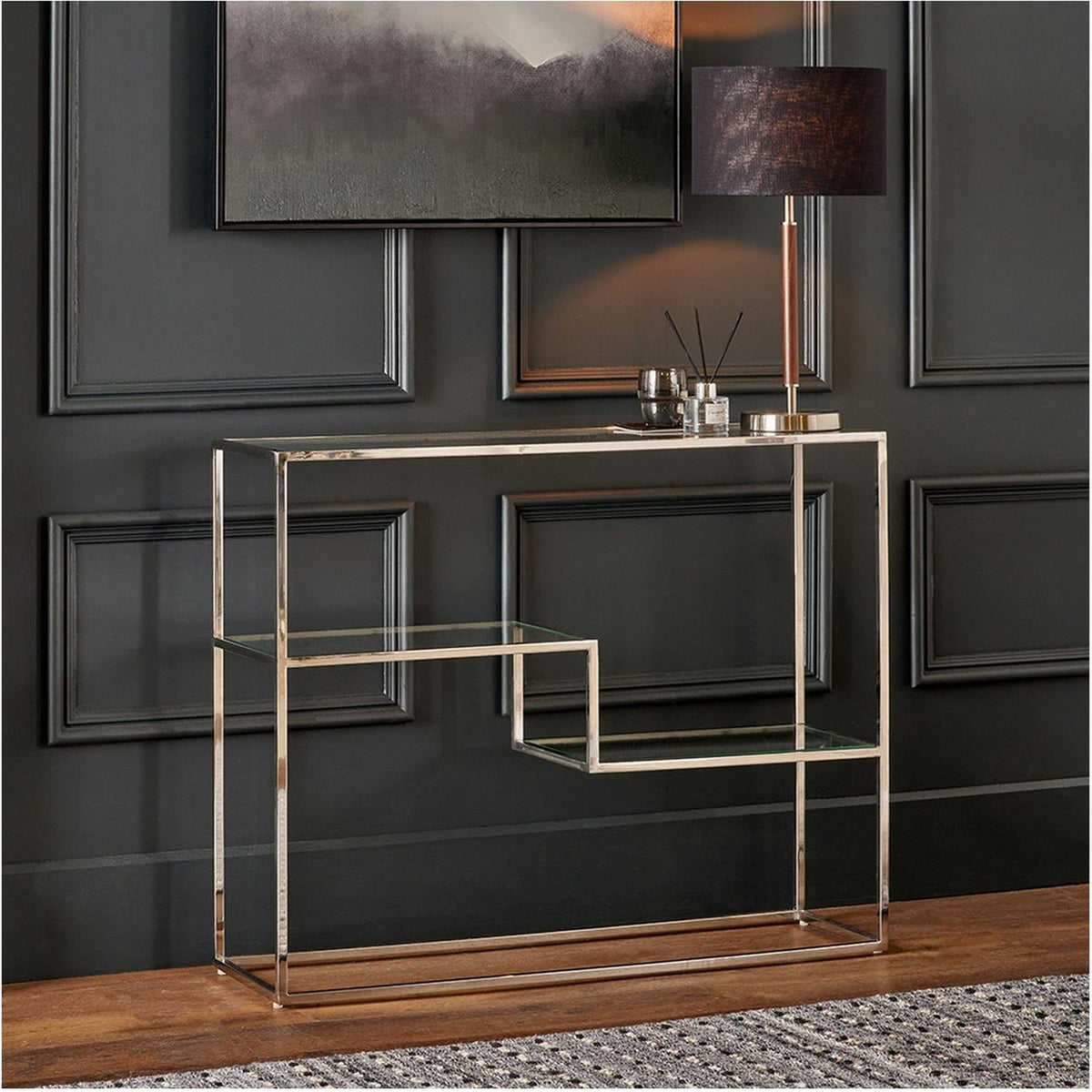 Aleandra console table with clear glass top and shelves on a chrome metal frame, styled with a lamp and decor against dark panelled walls.