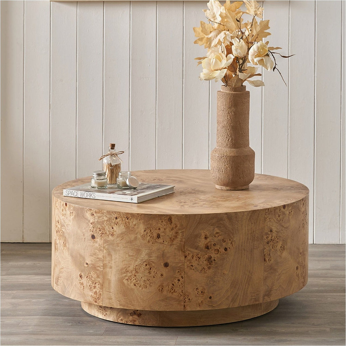 Round Newborough coffee table in burr wood effect finish with cylindrical base, styled with a textured vase, dried flowers, and books in a living room setting.