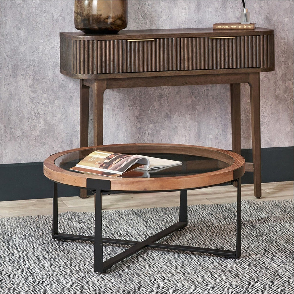 Vermont round coffee table with clear glass insert and light walnut wood veneer surround on a black metal cross-frame base, shown in a living room beneath a wooden console table.