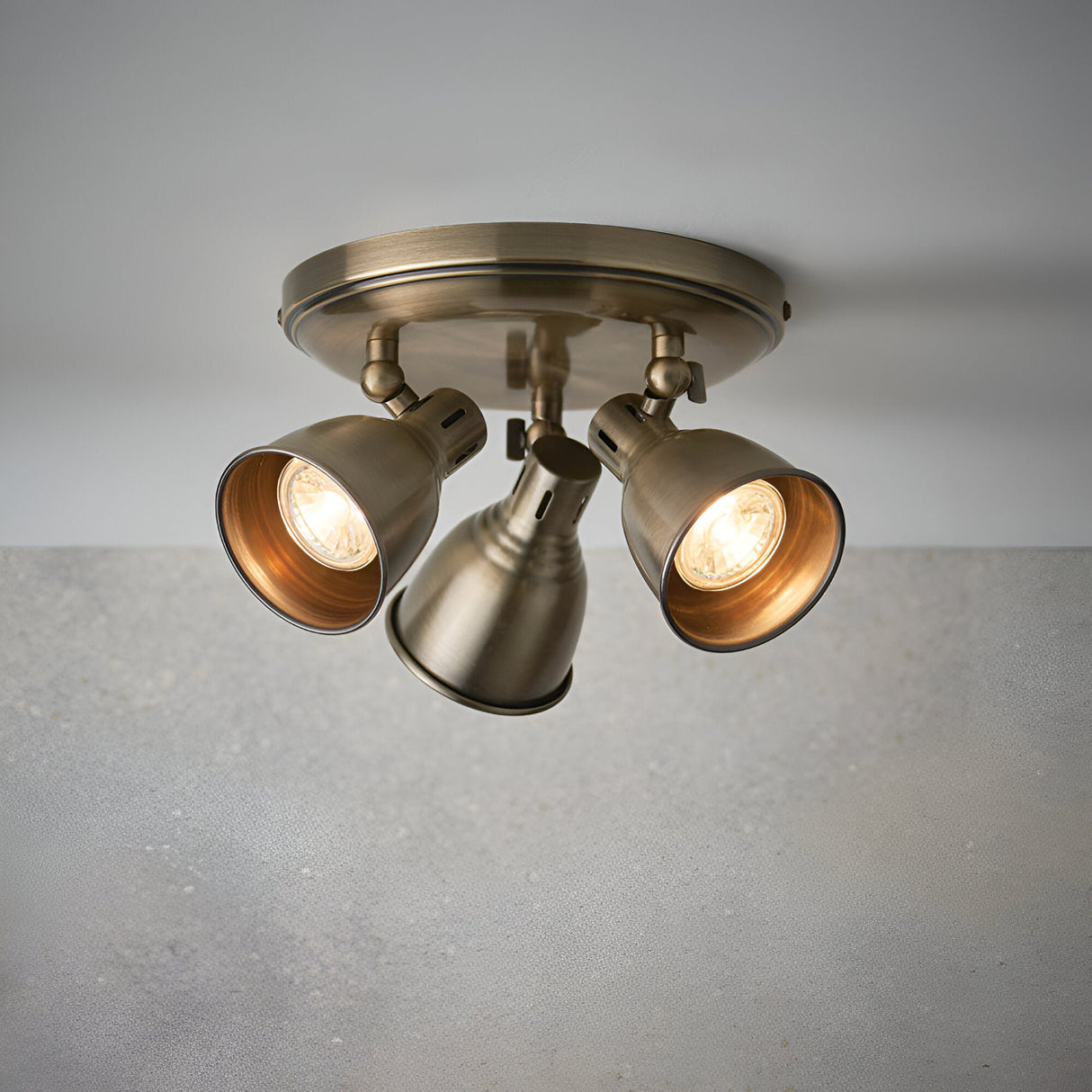 Endon Daneford 3 light round ceiling spotlight in antique brass with three independently adjustable steel heads, shown lit against a plain grey background.
