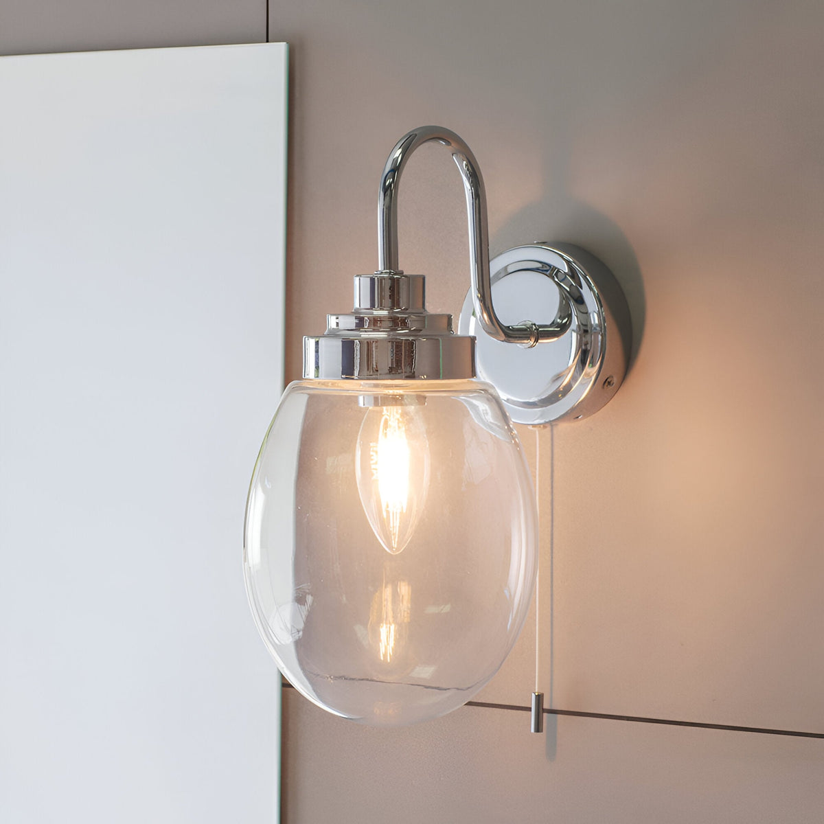 Endon Bramworth bathroom wall light in chrome with a curved arm, clear glass oval shade, pull cord switch, and E14 bulb, mounted beside a mirror.
