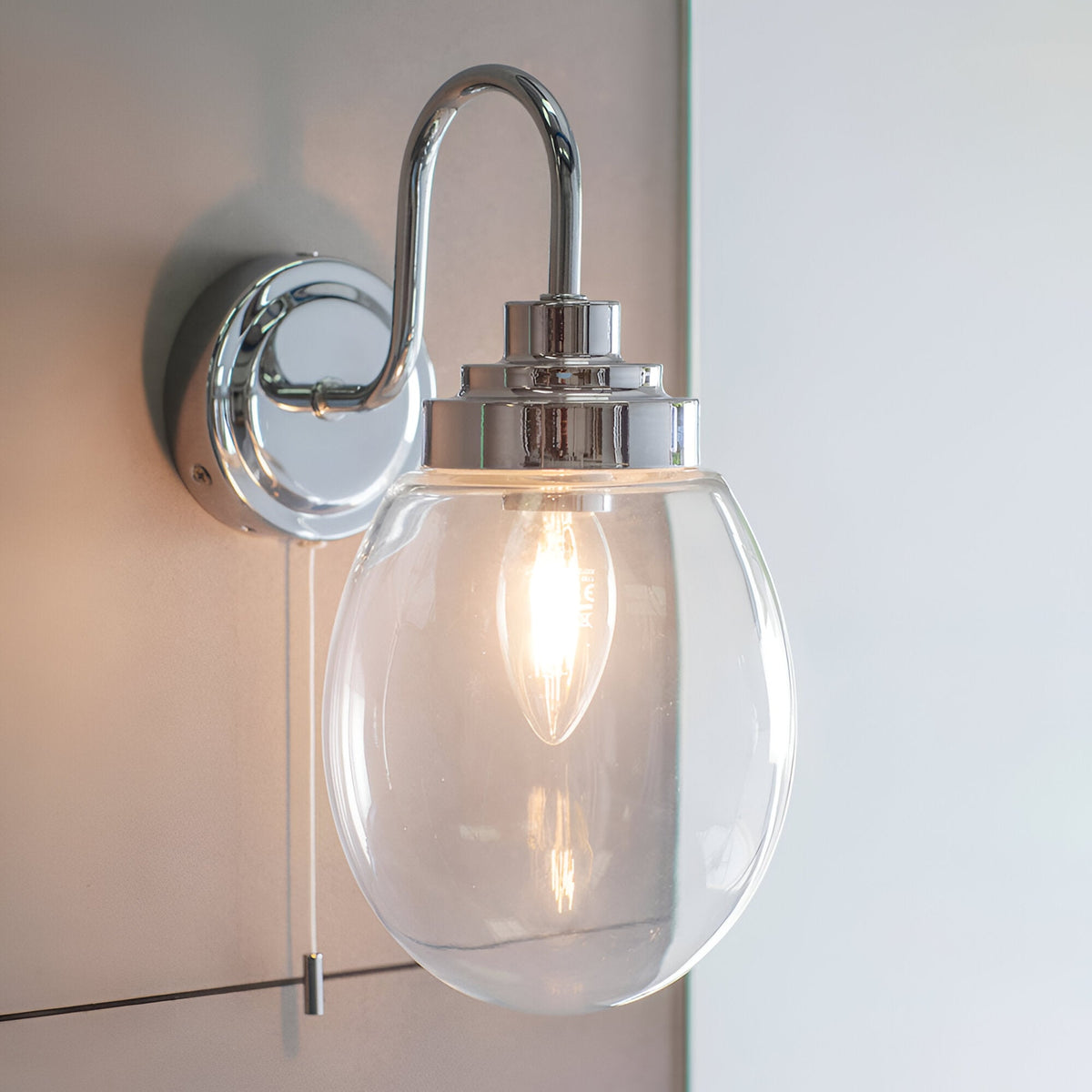Endon Bramworth bathroom wall light in chrome with a clear glass globe shade, arched arm, pull cord switch, and single E14 bulb, mounted beside a mirror.