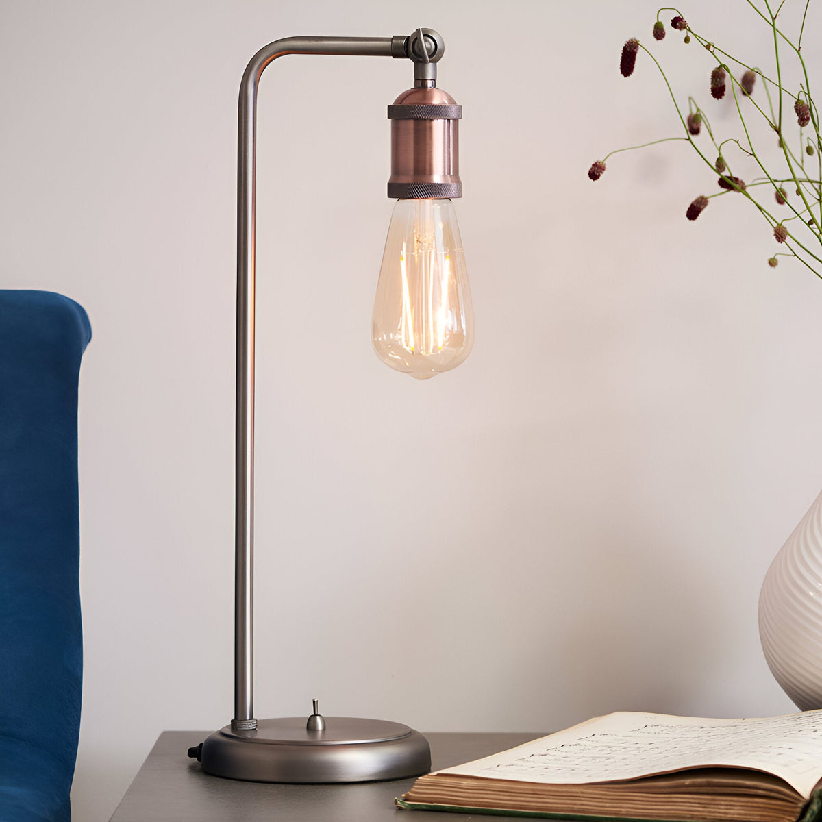 Brackley Table Lamp in silver and copper with an exposed E27 bulb, angled arm, and round base switch, shown on a bedside table.