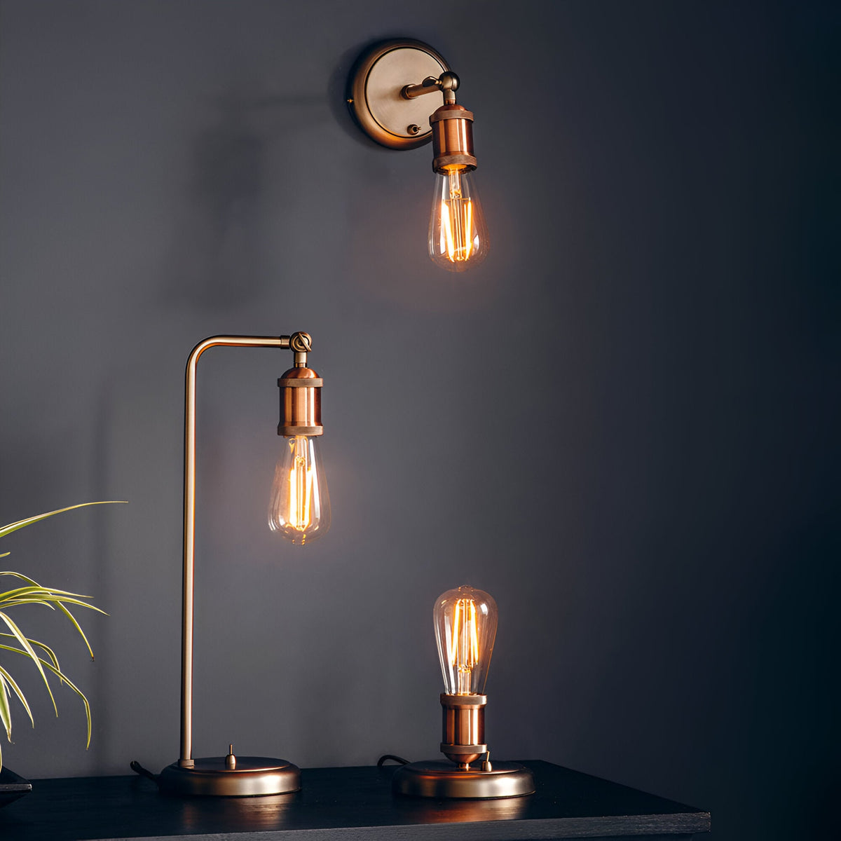 Brackley Table Lamp in copper with silver base, adjustable industrial-style arm and exposed E27 bulb, shown lit on a dark shelf beside a matching wall light.
