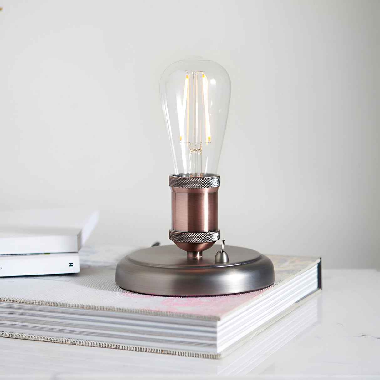 Endon Brackley industrial table lamp in aged pewter and copper with knurled E27 holder and exposed filament bulb, shown on books in a bright interior.