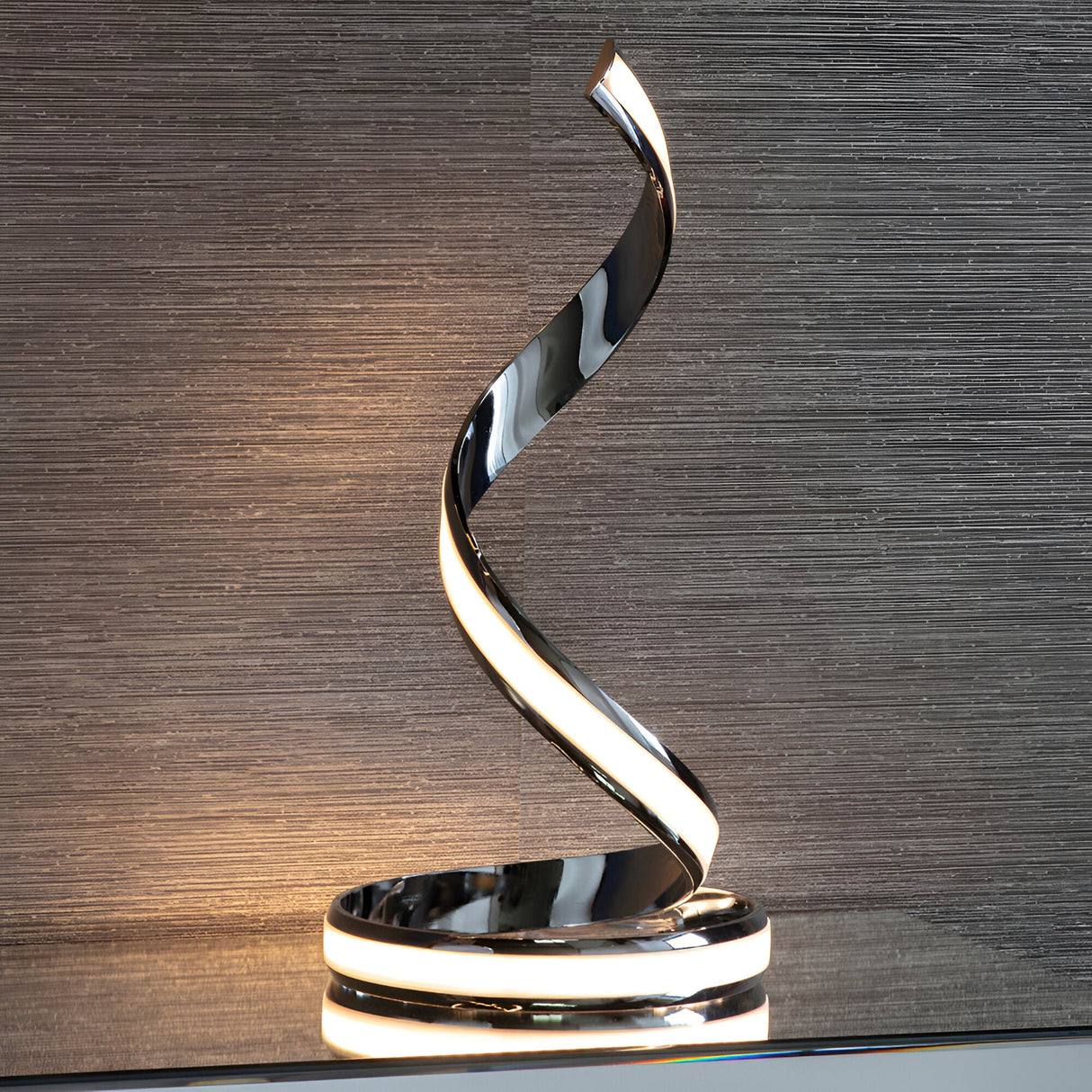 Endon Neve table lamp in polished chrome with a spiralling ribbon form and integrated LED diffuser, shown on a shelf against a textured grey wall.