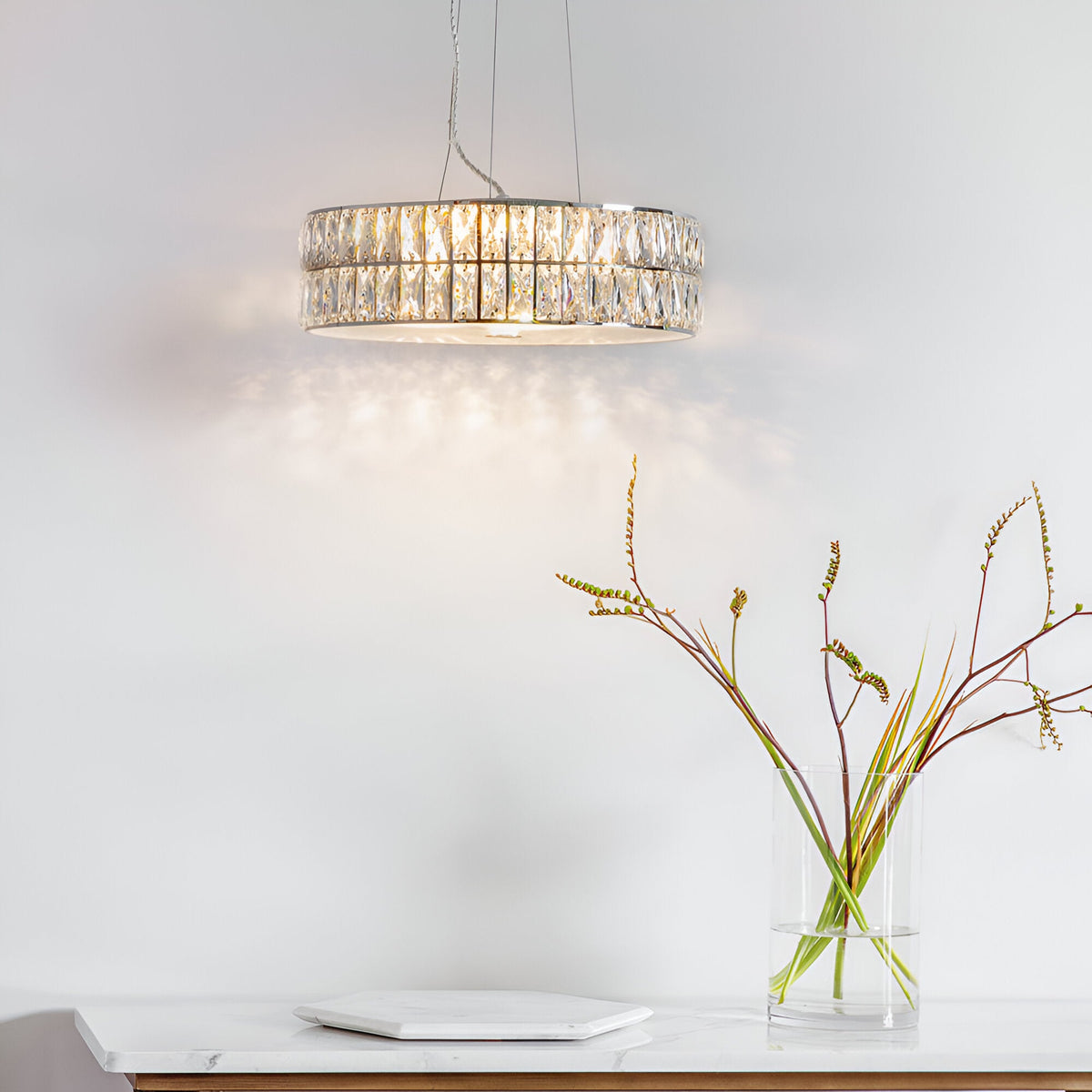 Marguerite LED round pendant in polished chrome with faceted crystal band, lit and hung above a marble console with vase and dish in a light room.