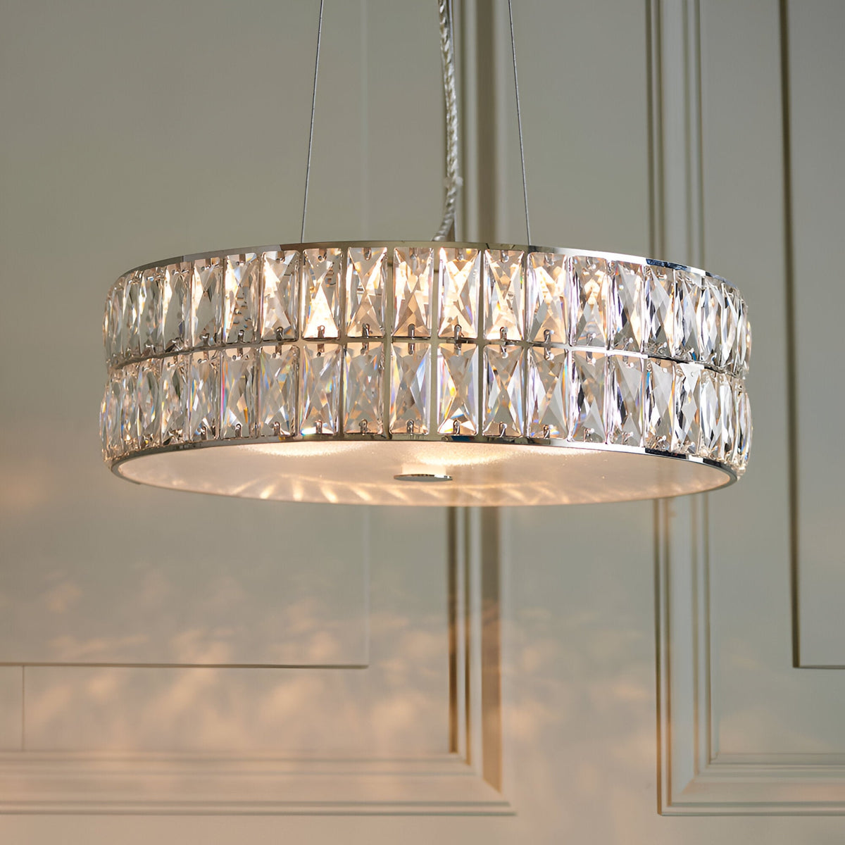 Marguerite LED round pendant in polished chrome with two tiers of faceted crystal panels, illuminated and suspended from clear cables in a panelled room.