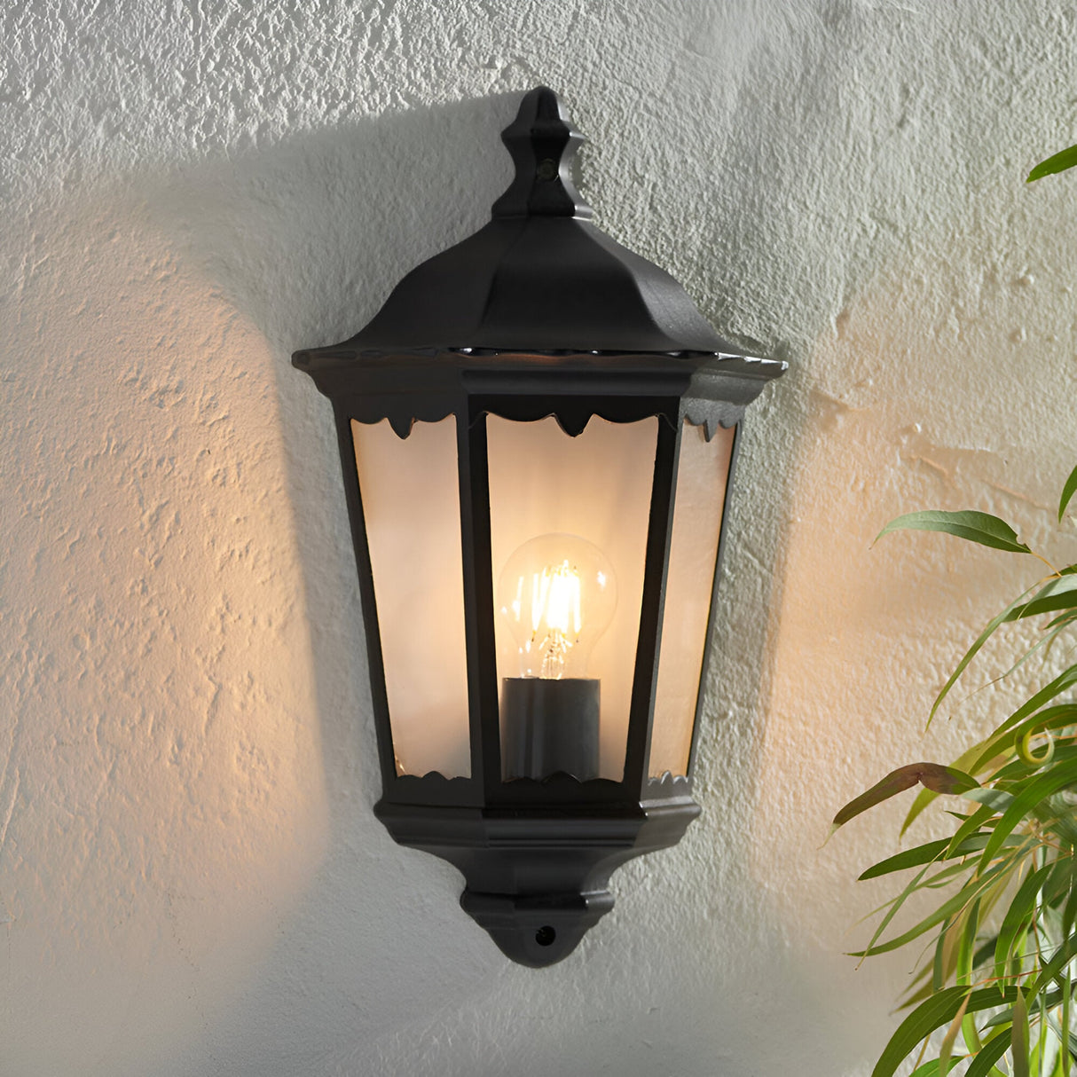 Ashshaw Coach Lantern in matt black die-cast aluminium with clear glass panels, mounted on a textured wall and lit with a visible bulb.