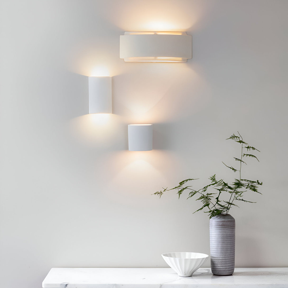 Endon Bramhurst white ceramic wall light illuminated alongside two smaller ceramic wall lights, above a marble shelf with a grey vase and white bowl.