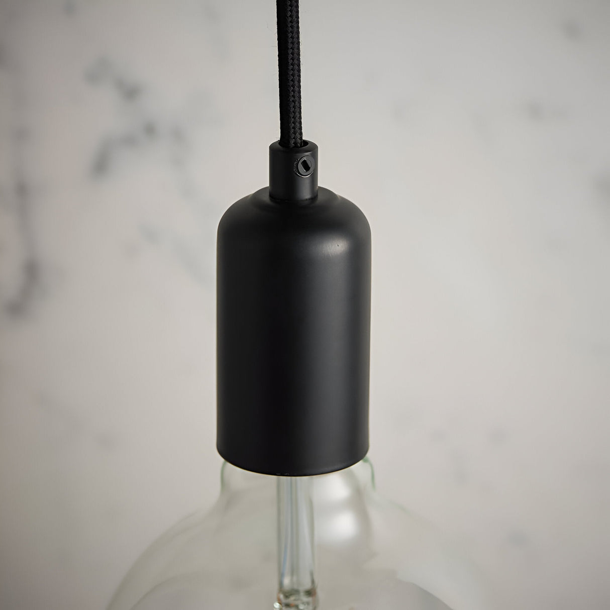 Close-up of the Endon Marlowe matt black E27 lampholder on black fabric flex, with a clear filament bulb below against a marble backdrop.