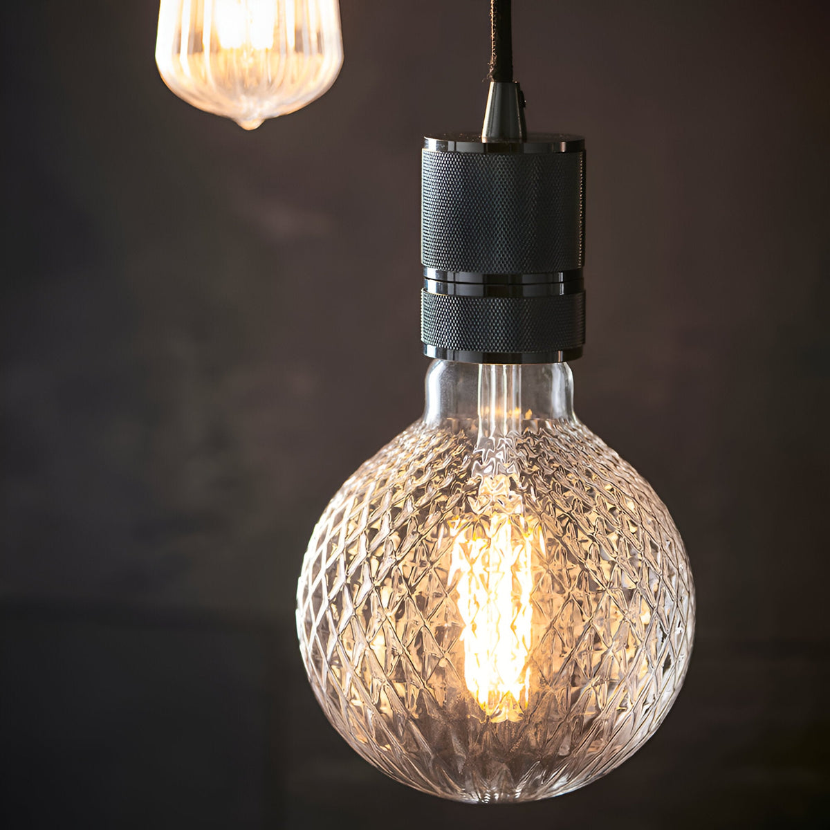 Endon Camden pendant light with knurled black chrome cable fitting and exposed textured glass globe filament bulb, second pendant glowing in the background.