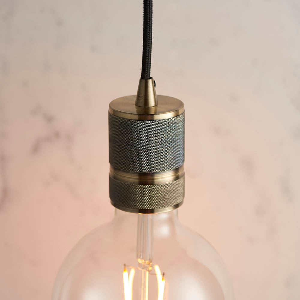 Endon Camden pendant light in antique brass with knurled lamp holder, black fabric flex and exposed E27 filament bulb, against a soft marble backdrop.