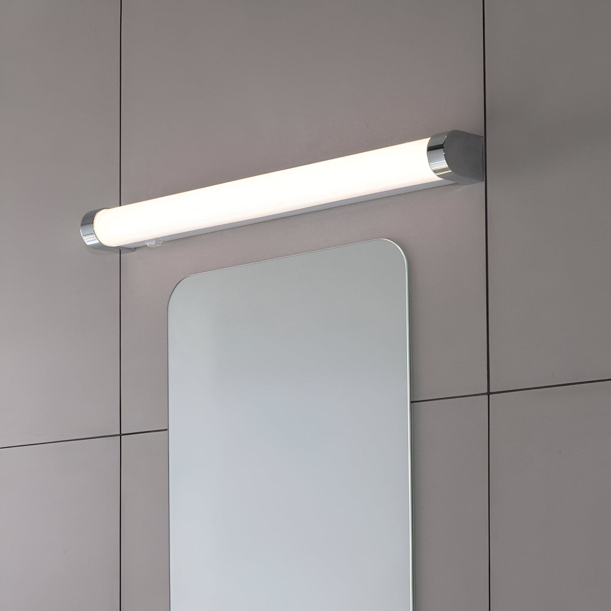 Endon Carlshaw large LED bathroom wall light with slim white ribbed diffuser and chrome end caps, mounted above a rounded rectangular mirror on grey tiles.