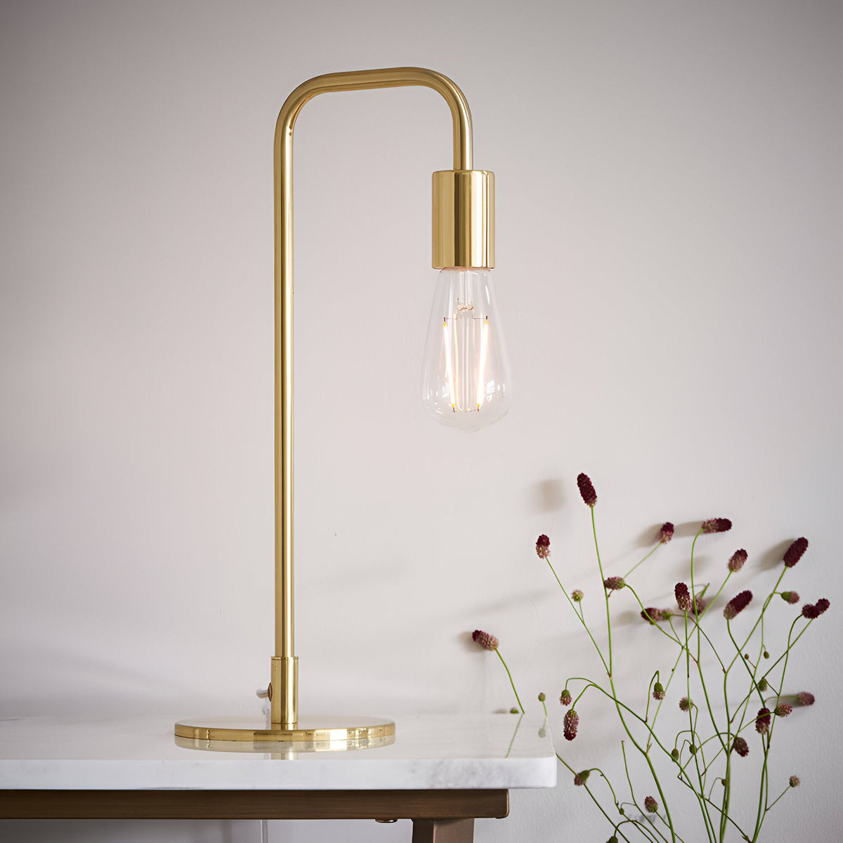 Endon Cliffgrove brushed brass table lamp with slim arched frame and exposed filament bulb, shown on a marble surface beside dried flowers.