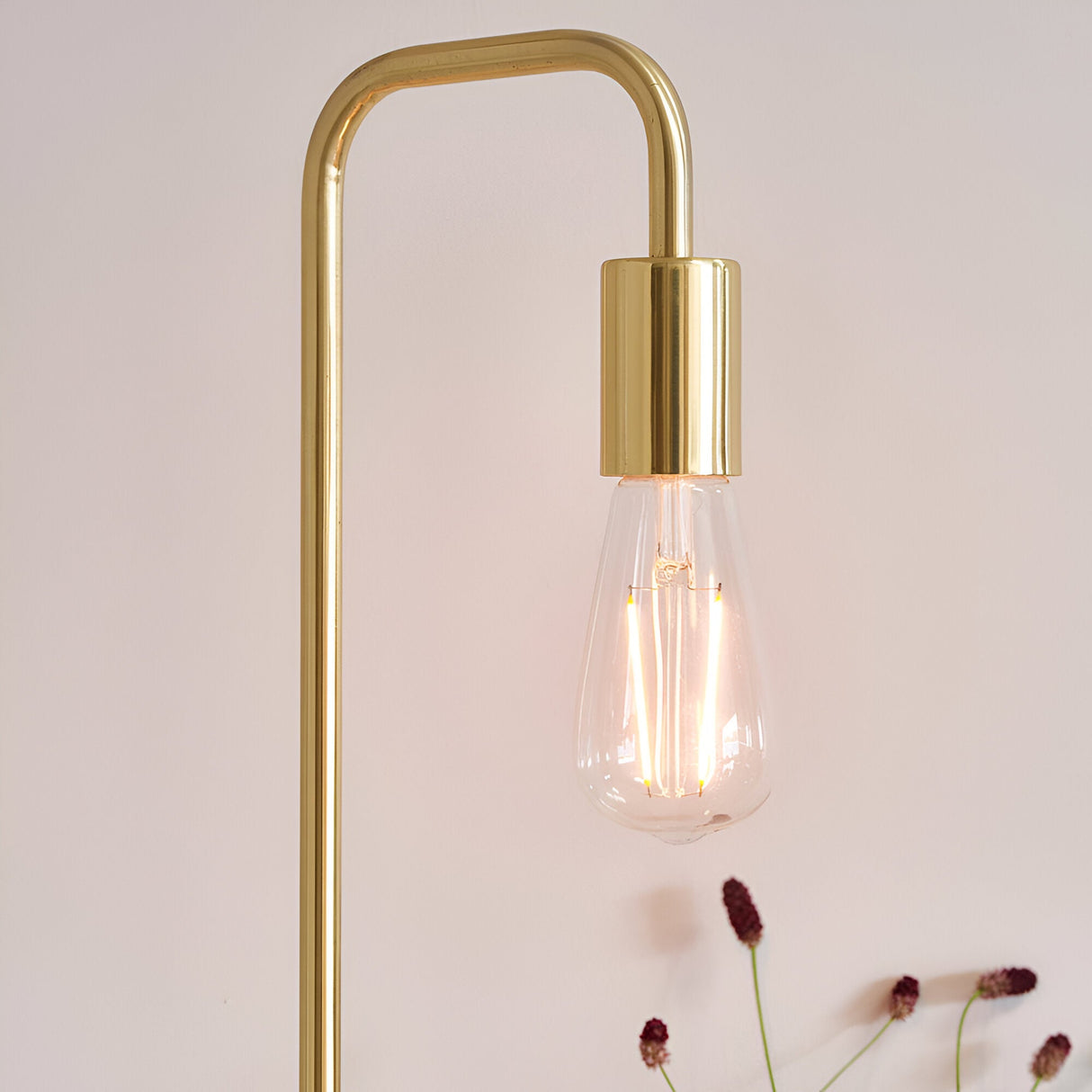 Endon Cliffgrove table lamp close-up showing slim brushed brass frame with exposed E27 filament bulb glowing against a soft neutral background.