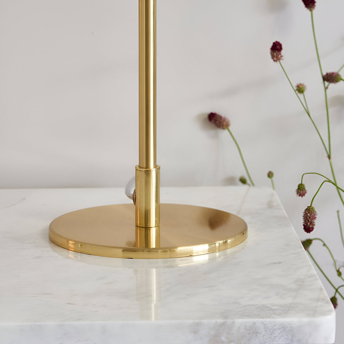 Close-up of the Endon Cliffgrove table lamp's round brushed brass base and slim stem on a marble surface beside dried pink flowers.